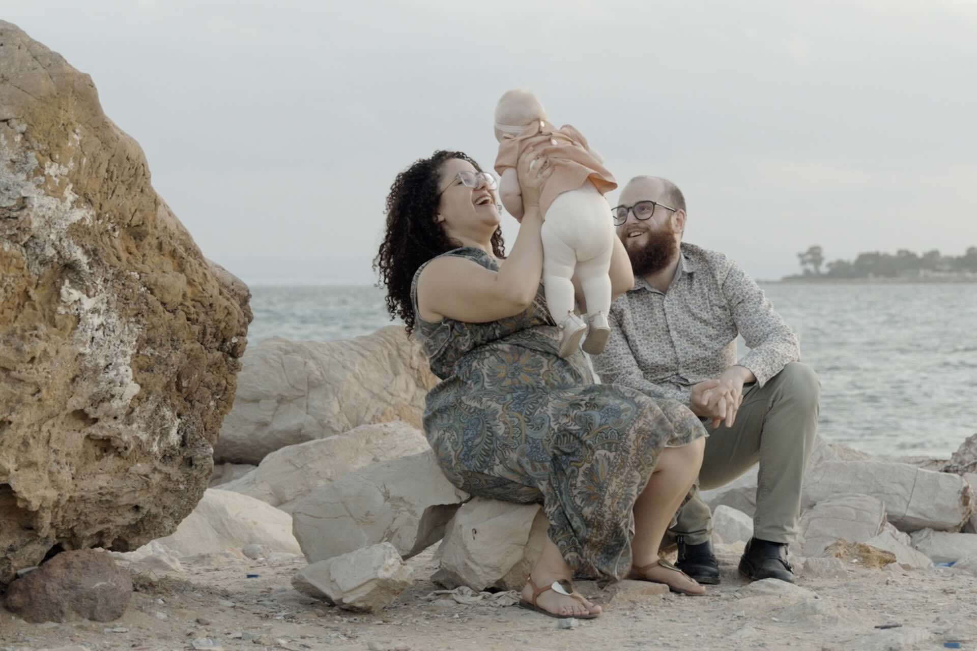 Parents laughing with their baby while sitting on rocks by the sea